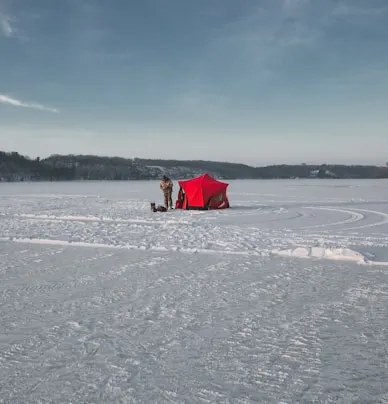 An ice fishing tent set up in the middle of a snowy lake, with a person fishing outside.